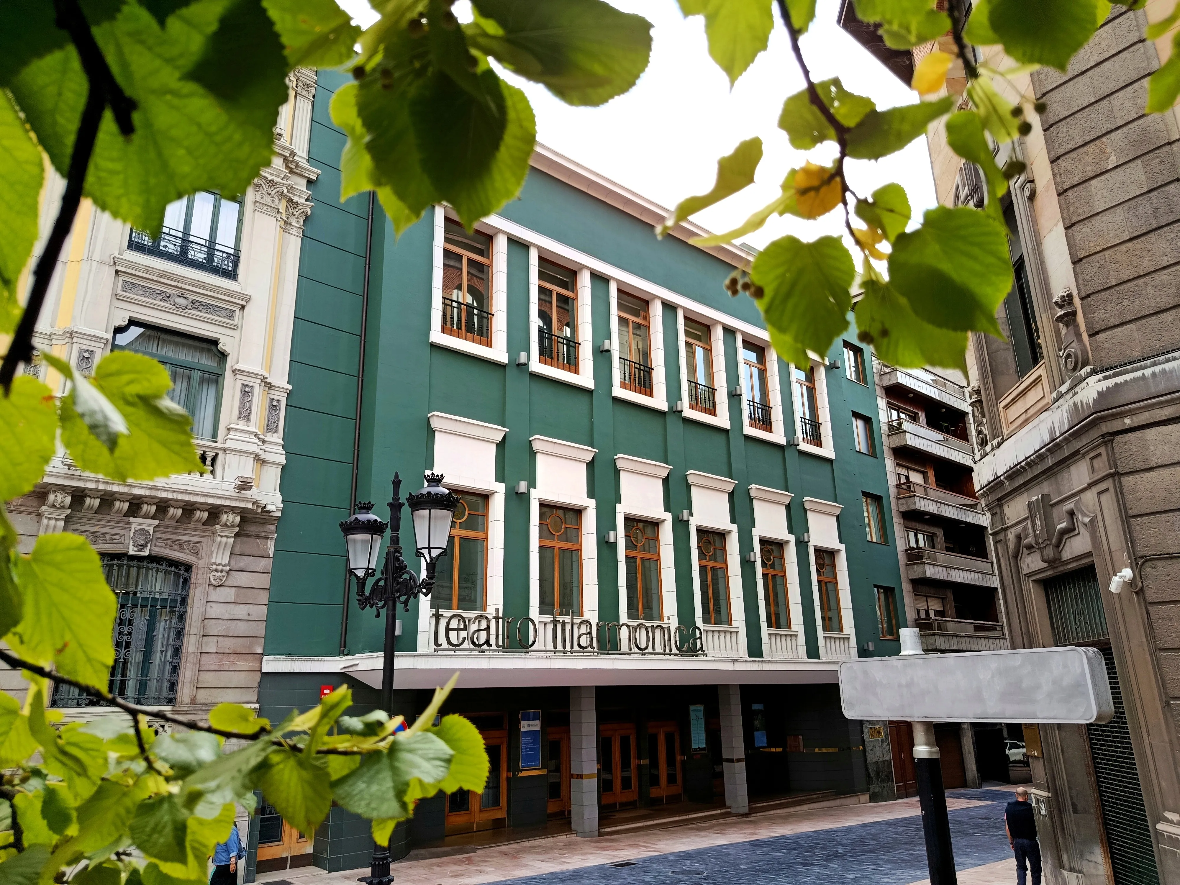 Fotografía de la fachada del teatro Filarmónica, emarcada por hojas de árbol